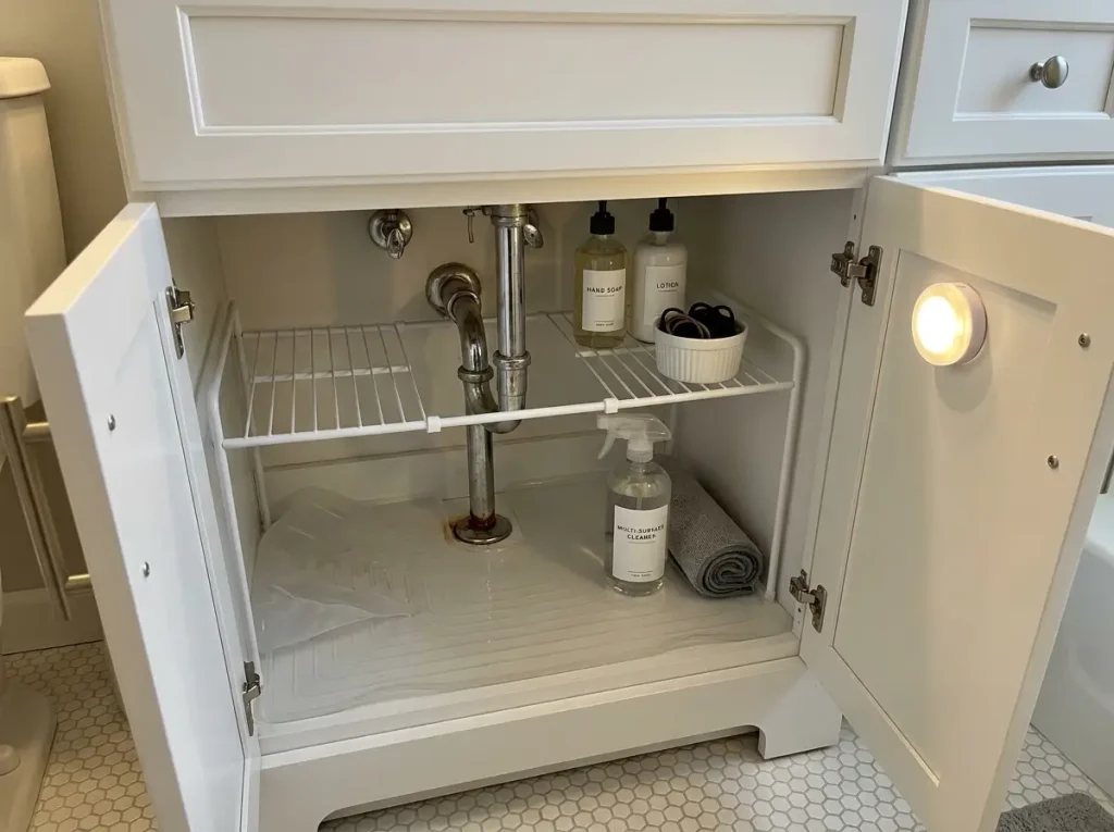 Open bathroom vanity cabinet showing two-tier expandable organizer around pipe with drip mat