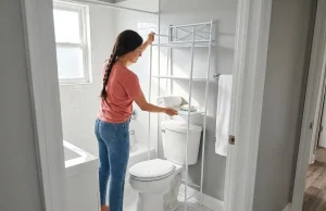 I Tested 6 Over-Toilet Shelves and This One Held Up Woman measuring over-toilet storage shelf in small white bathroom