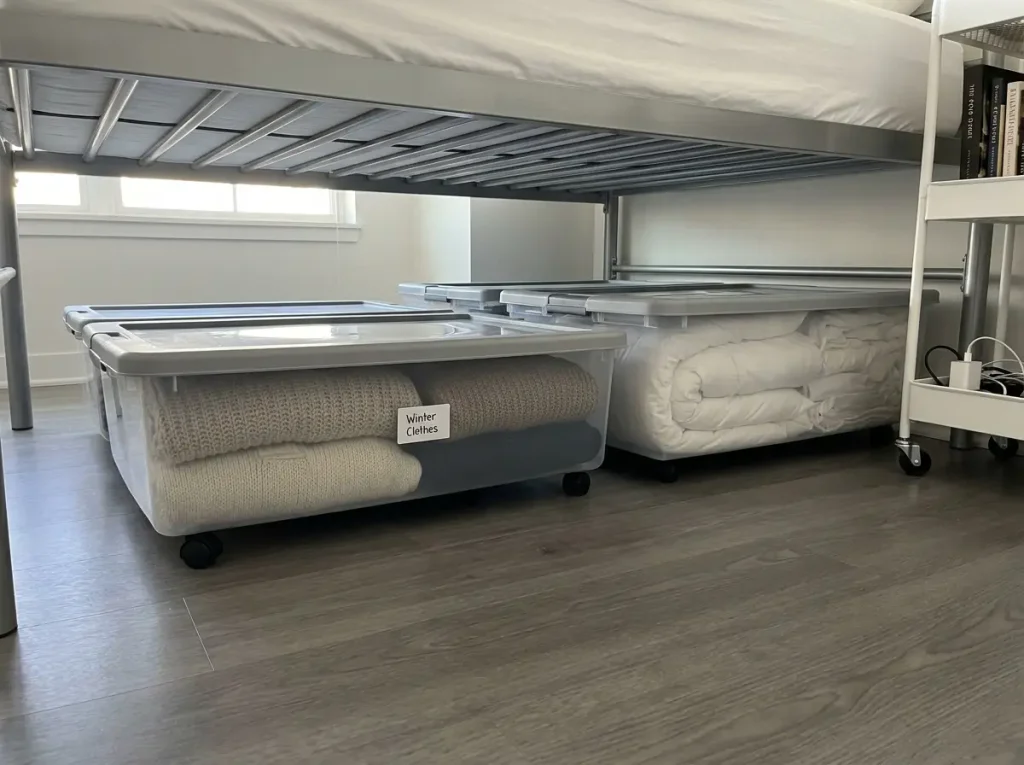 Organized under-bed storage containers on wheels visible beneath metal bed frame in small apartment bedroom