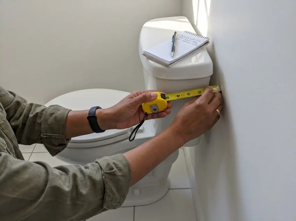 Hands measuring the gap between toilet tank and wall with a tape measure before buying over-toilet storage