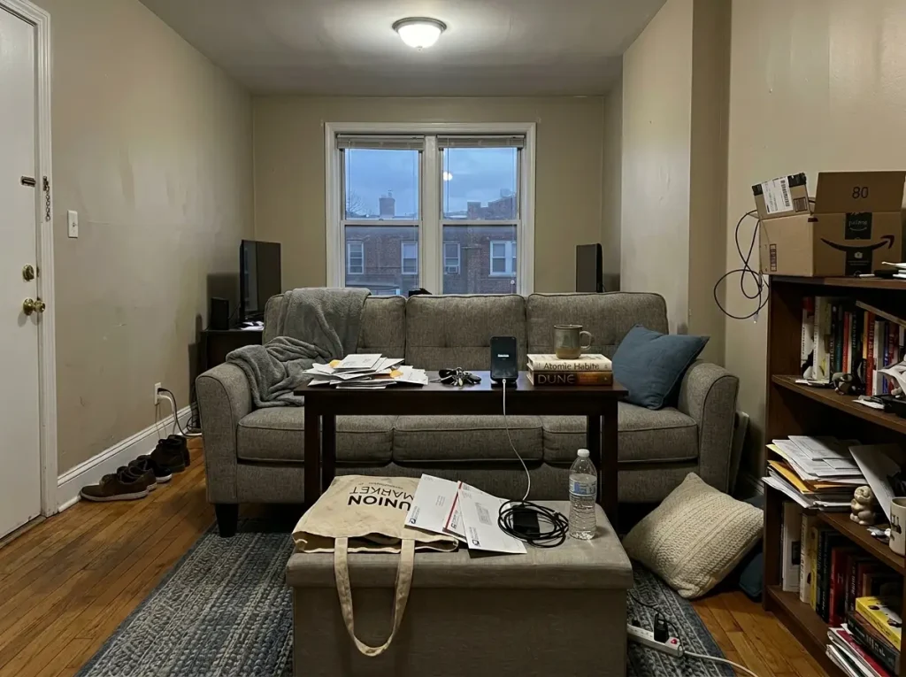 Small apartment living room showing drift — accumulated objects on every surface after weeks of use