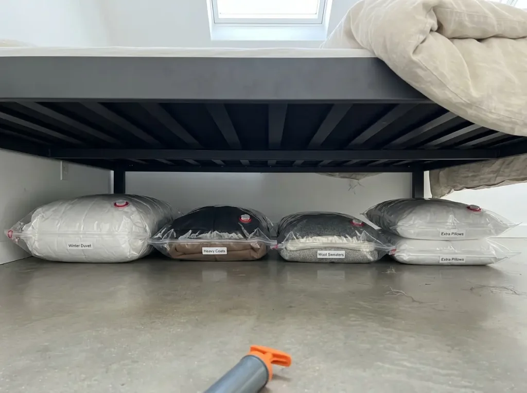 Four vacuum storage bags stacked flat under low platform bed showing compressed duvet and winter coats through clear panels