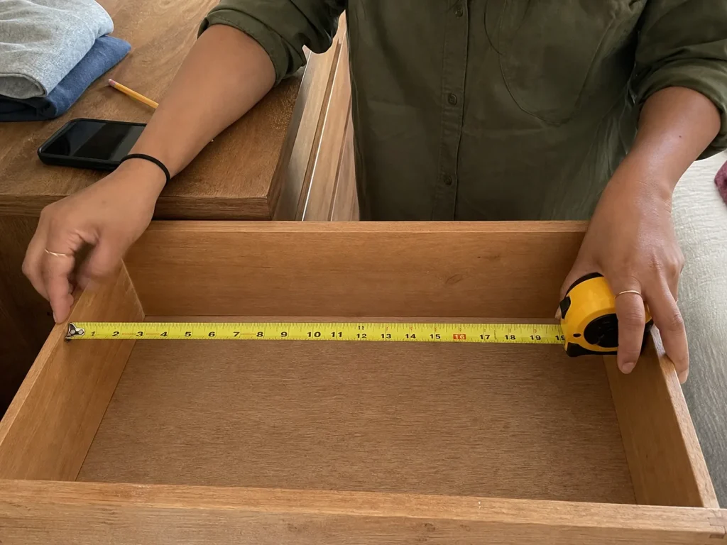 Hands measuring the interior width of an open dresser drawer with a yellow tape measure