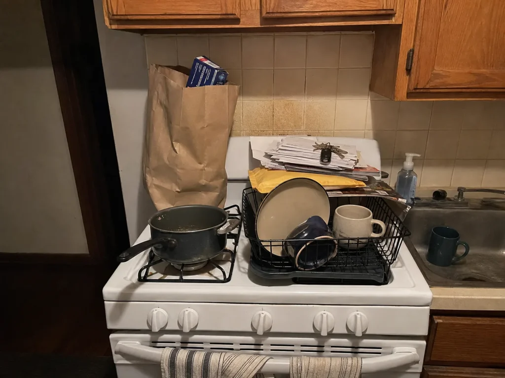 Mail, dishes and groceries piled on a stovetop in a kitchen with no counter space