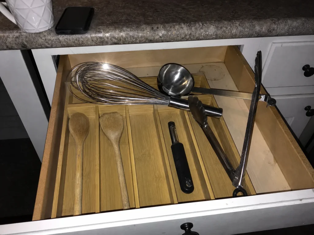 A whisk sitting crosswise on a shallow flatware tray, jamming a kitchen utensil drawer