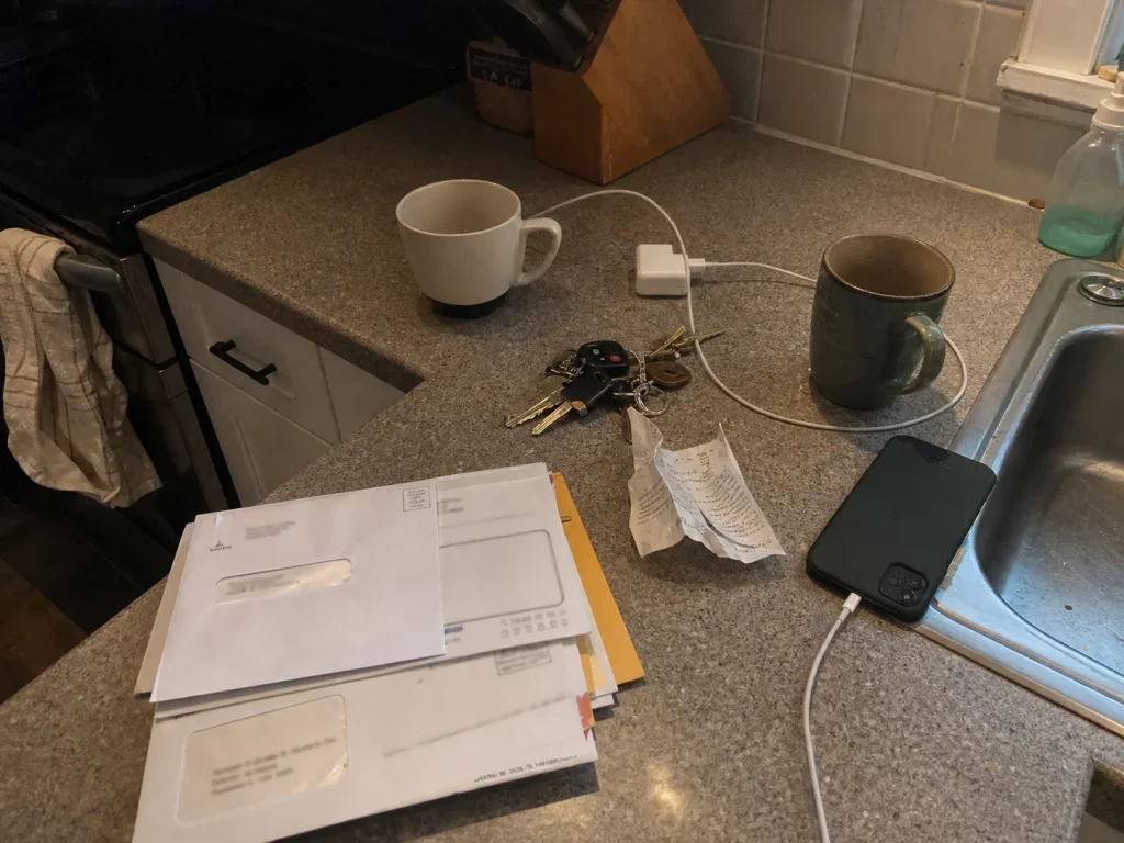 Kitchen counter mid-clutter with mail, keys, charger and mugs drifting onto the prep zone