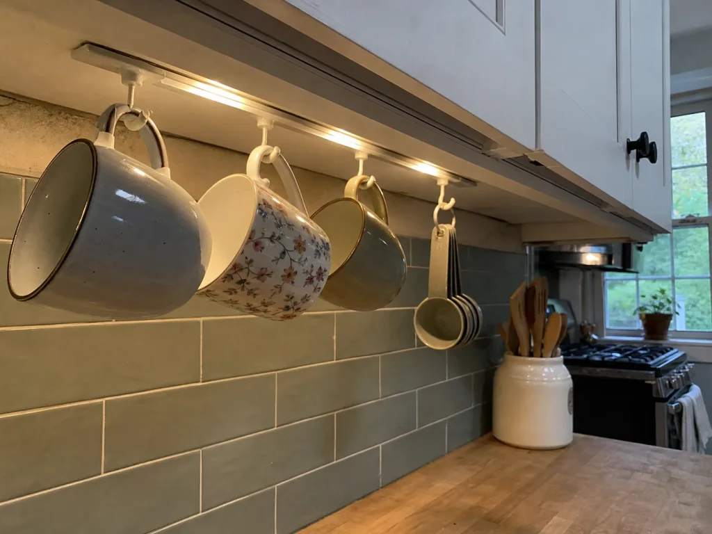 Mugs and measuring cups hanging from adhesive hooks under an upper kitchen cabinet to free counter space
