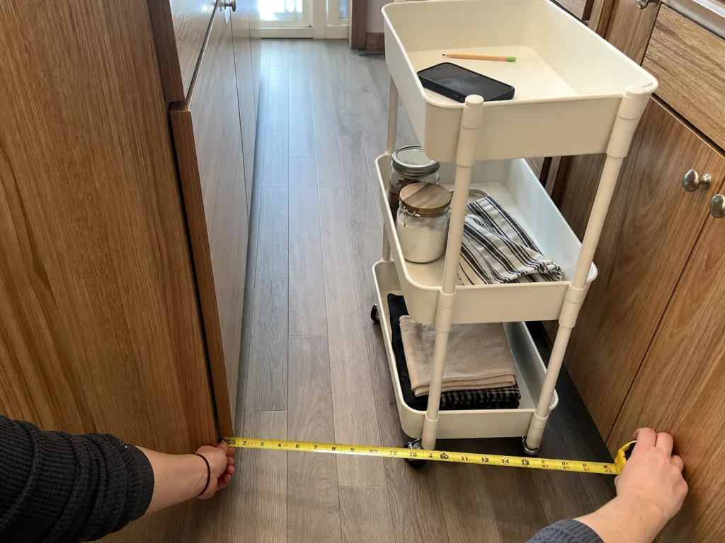 Hands measuring the aisle gap next to a slim rolling cart in a narrow galley kitchen