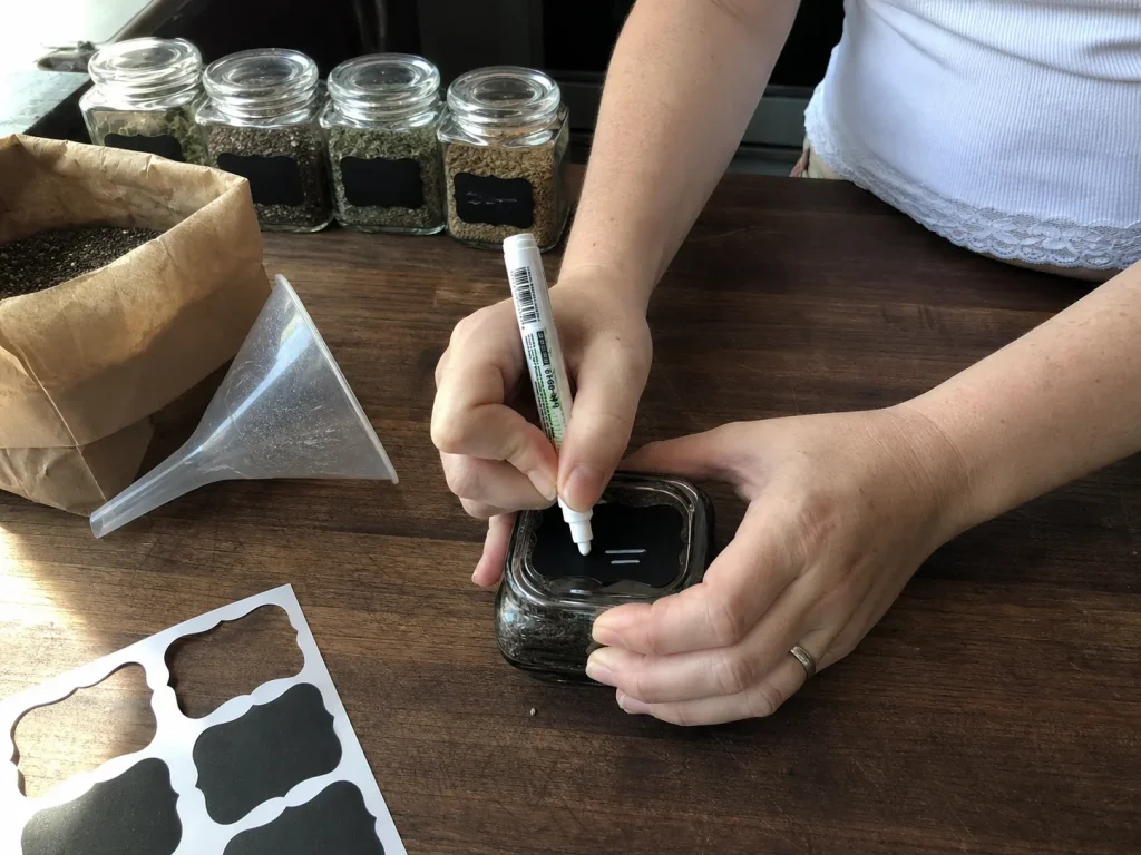 Spice Drawer Organization That Stops Jars From Rolling 12 Hands writing on a removable chalkboard label on a spice jar lid during a restock at the kitchen counter