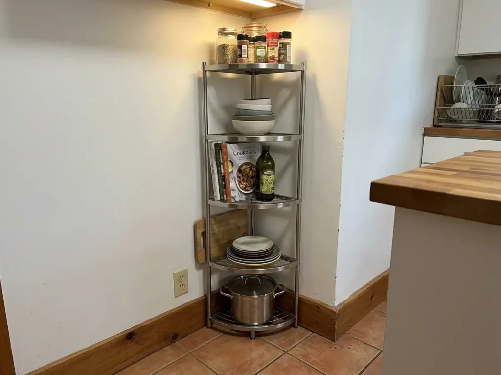 Freestanding stainless corner rack in a kitchen corner blocked by a baseboard and outlet