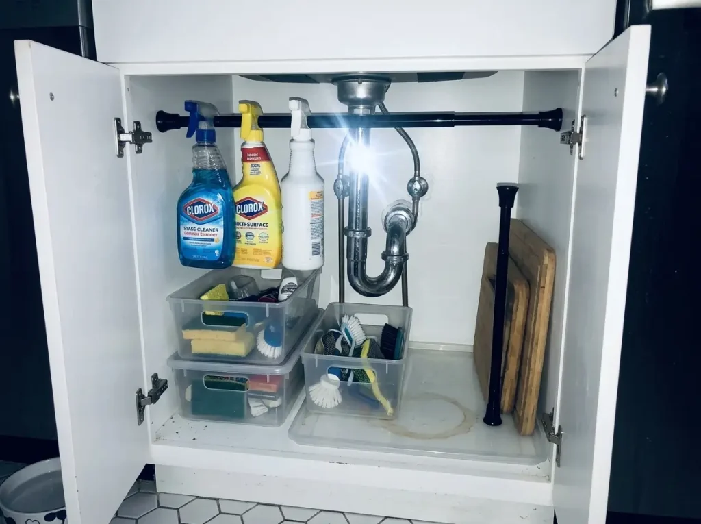 Under-sink cabinet with a small tension rod holding spray bottles above bins, lit by camera flash