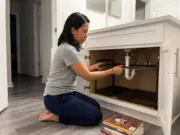 Best Under Sink Organizers for Bathroom Cabinets Woman kneeling at an open bathroom vanity fitting an expandable under sink organizer around the pipe