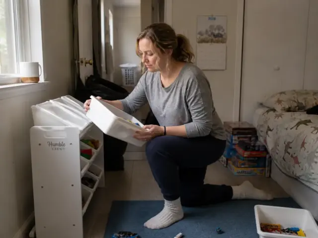 Mother sorting toys into a three-tier bin organizer in a small kids bedroom with toy storage on the wall