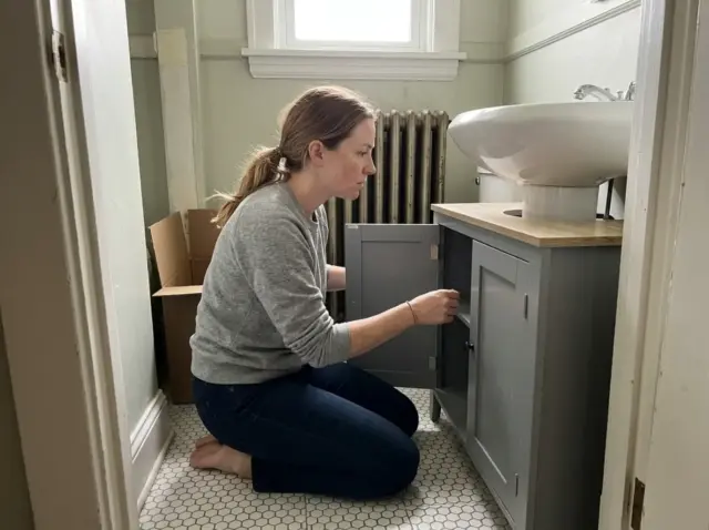 Woman kneeling beside a pedestal sink with a wrap-around under sink storage cabinet in an older bathroom