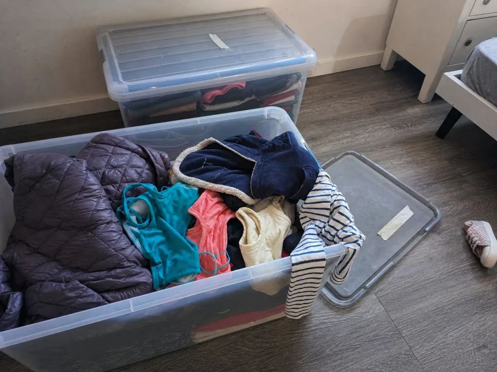 Open storage bin labeled size 5 with mismatched winter and summer kids clothes crammed together