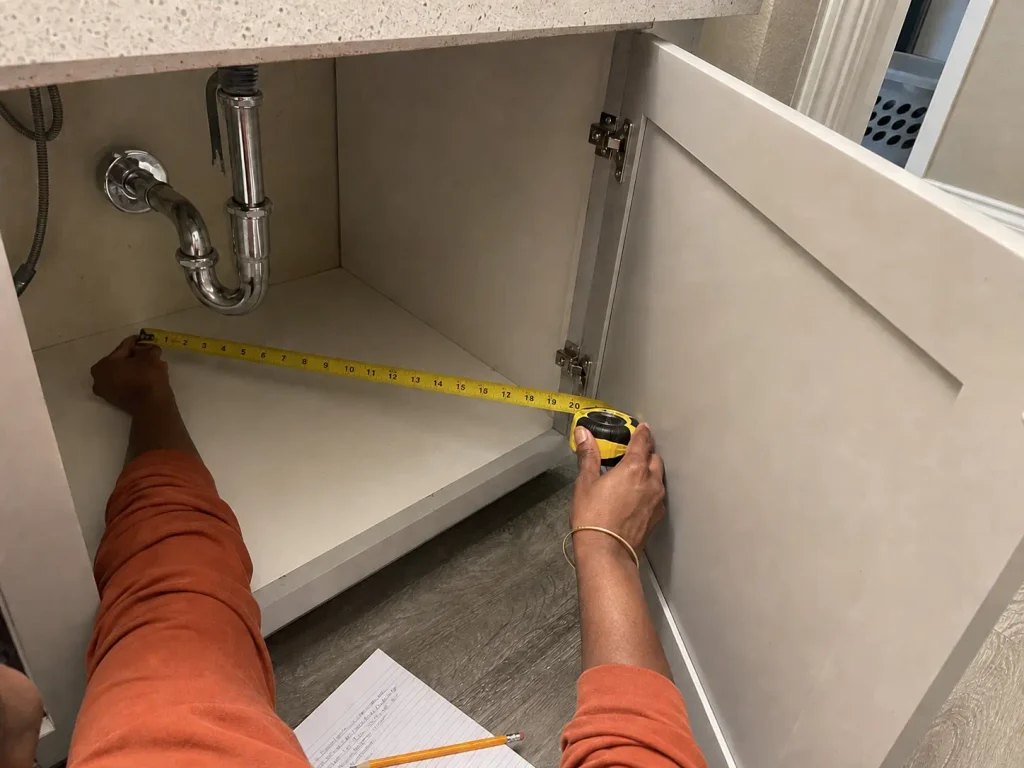 Hands measuring the interior depth of a bathroom vanity cabinet with a yellow tape measure beside the P-trap