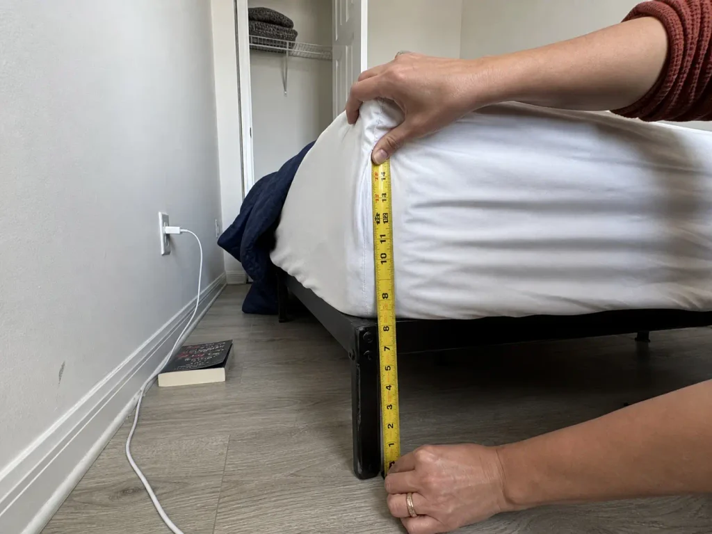 Tape measure held from the floor to the mattress top to check nightstand height in a small bedroom