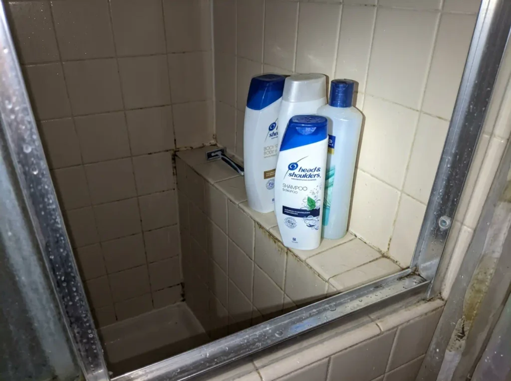 Crowded small shower ledge with bottles pushed to the edge in a narrow tiled stall