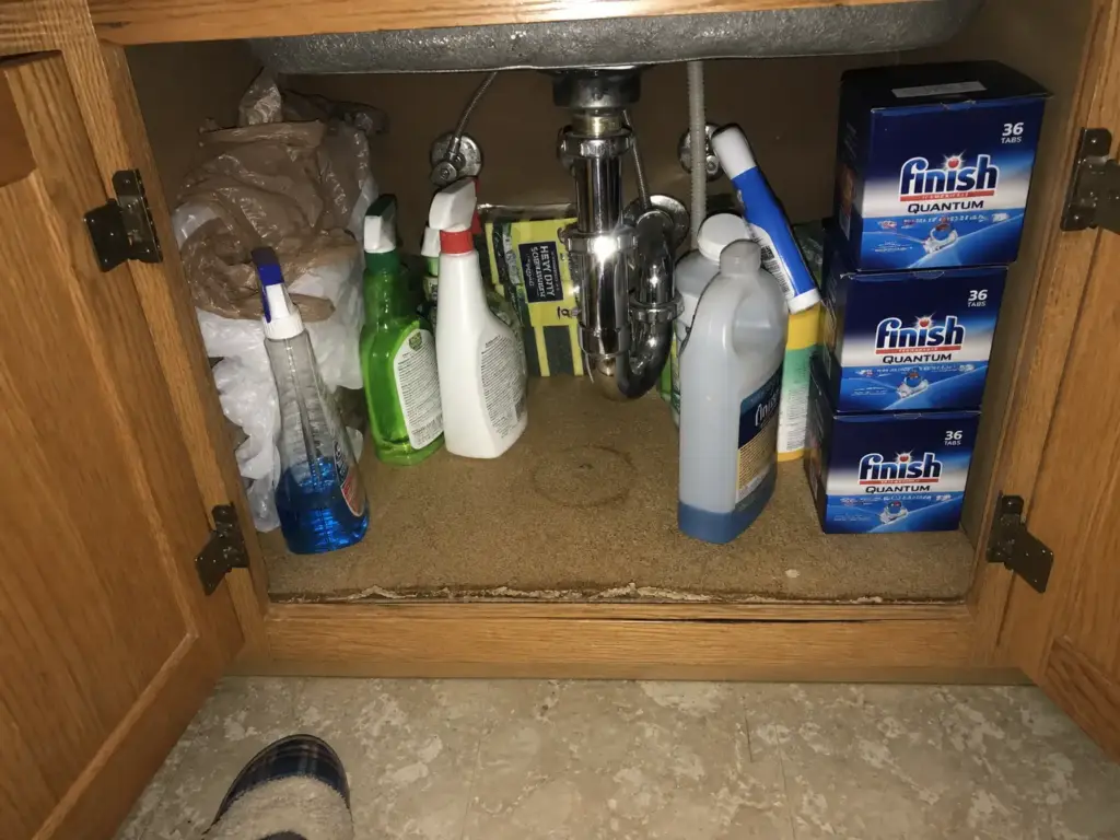 Cluttered under sink kitchen cabinet with bottles crowded around the P-trap and a damp cabinet floor