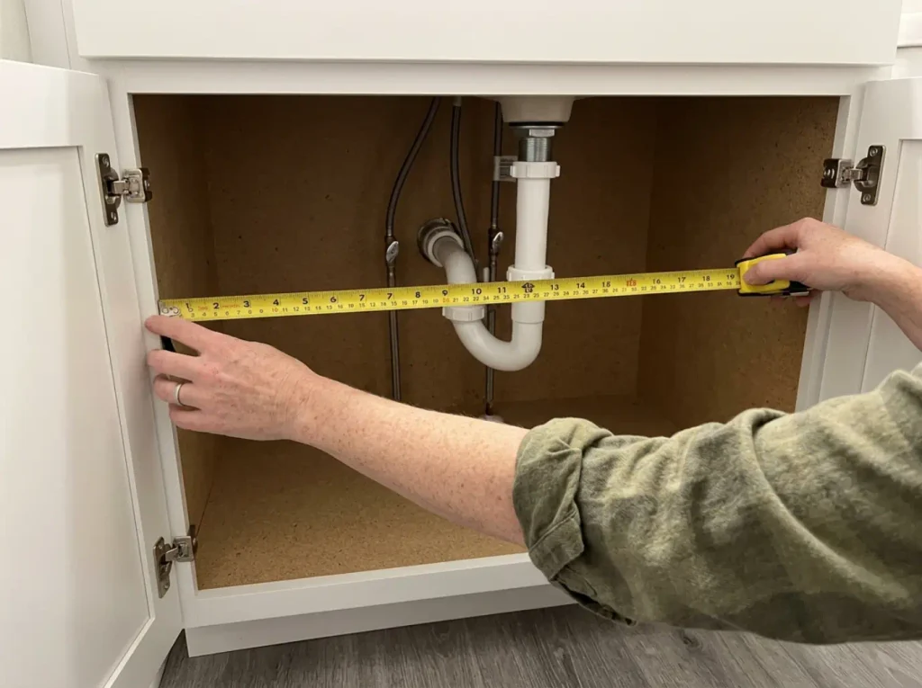 Tape measure held across the clear span beside a bathroom sink P-trap inside an empty vanity cabinet