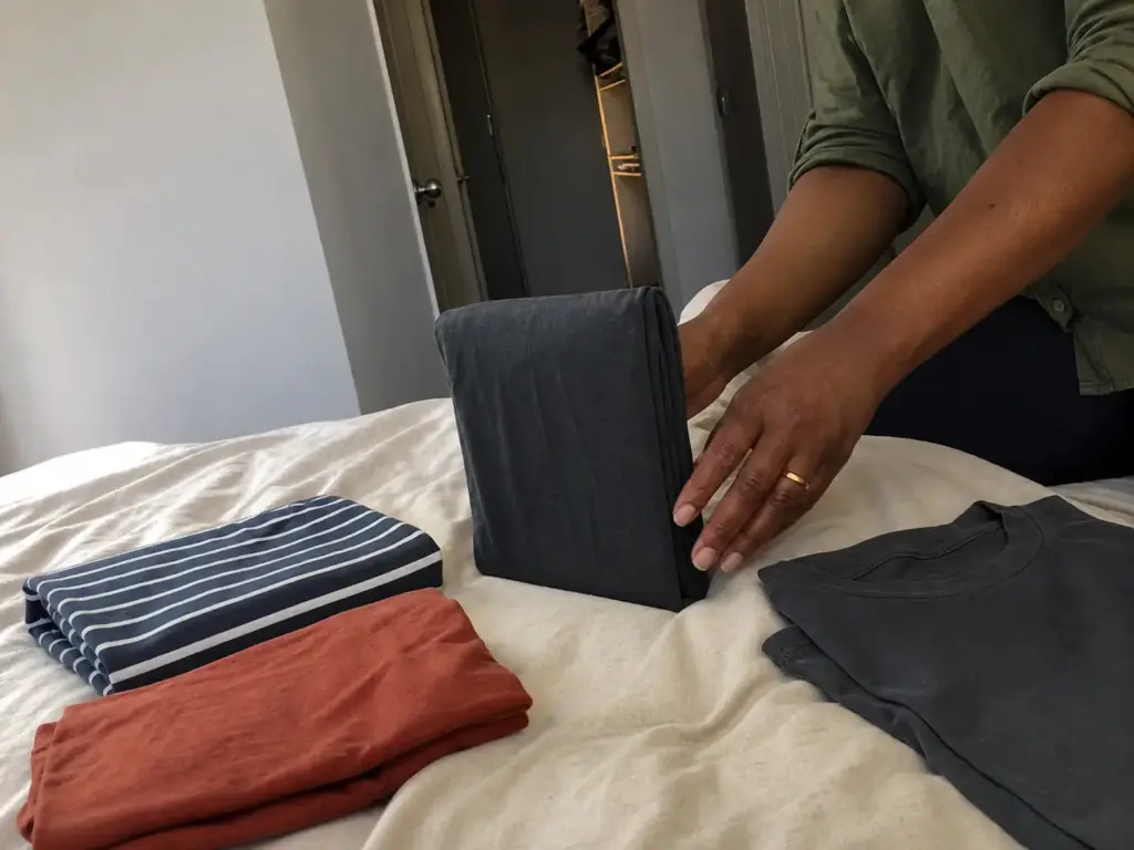 Close-up of hands folding a t-shirt into a compact rectangle that stands upright on a bed