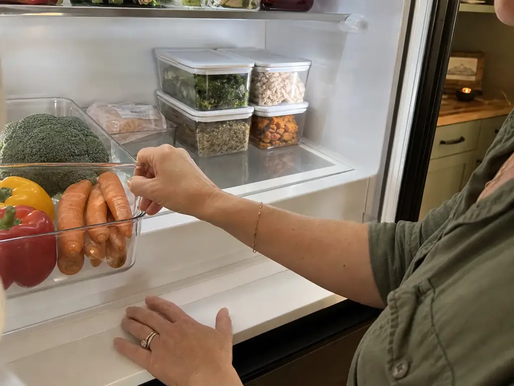 Hands setting a clear bin to divide a fridge shelf into raw-staged and ready-to-eat zones