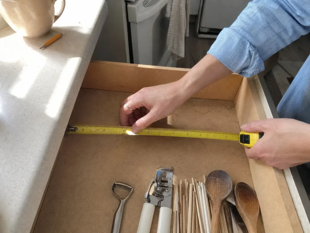 Why Your Small Kitchen With Few Drawers Stays Cluttered 2 Hands measuring the interior width of a kitchen drawer with a yellow tape measure before buying dividers