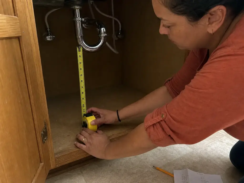 Hands measuring vertical pipe clearance under a kitchen sink with a yellow tape measure