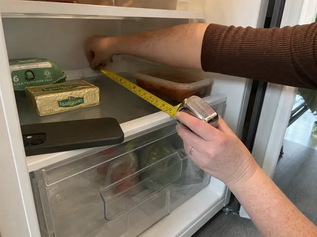 Hands measuring the shelf depth of a small fridge with a tape measure before buying bins