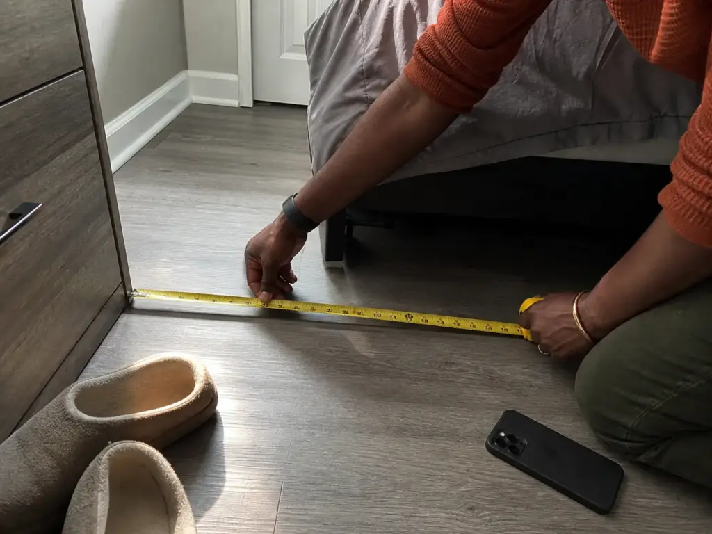 Hands measuring the walkway clearance beside a bed with a yellow tape measure