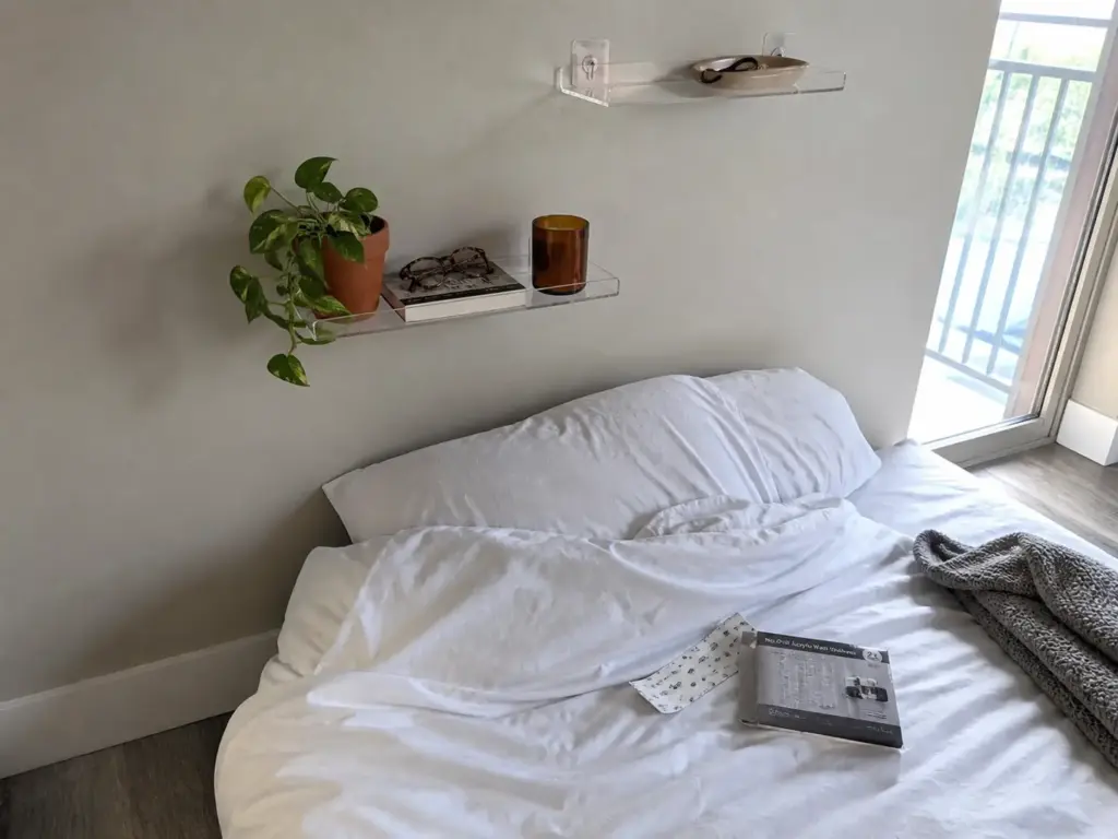 Clear no-drill acrylic wall shelf mounted above a bed holding a book, glasses and a small plant