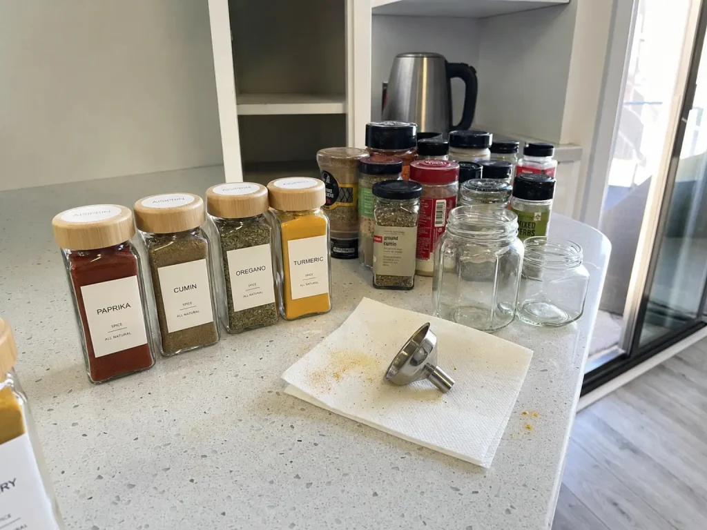 Matching glass spice jars with bamboo lids lined up beside leftover original store bottles on a counter