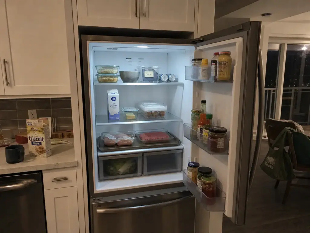 Open refrigerator organized by zones with leftovers on top, dairy in the middle and raw meat on a tray below