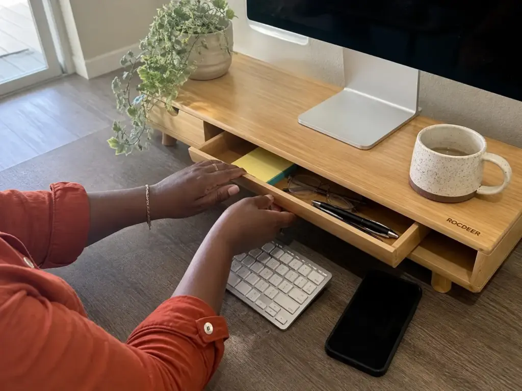 Aesthetic Desk Organization Ideas That Survive Week Two 5 Hands opening the drawer of a bamboo monitor riser holding desk supplies above a small desk