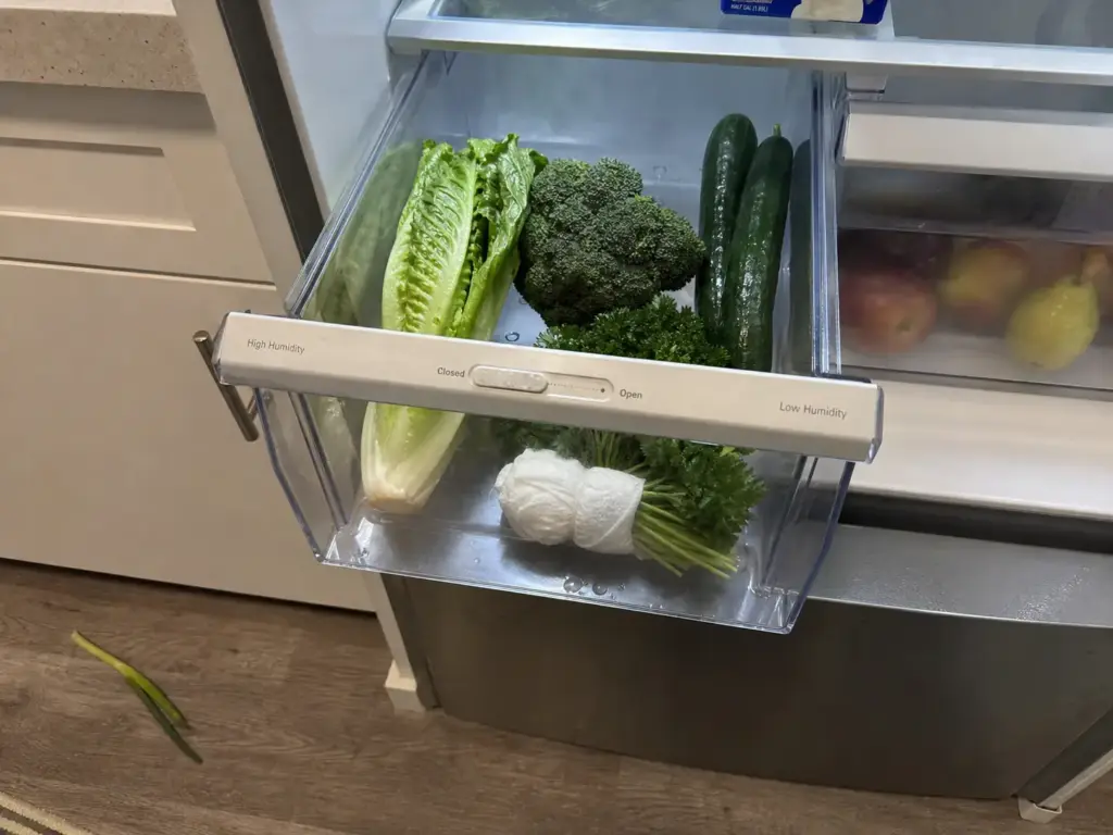 Open fridge crisper drawer with leafy greens and the humidity slider visible on the drawer front