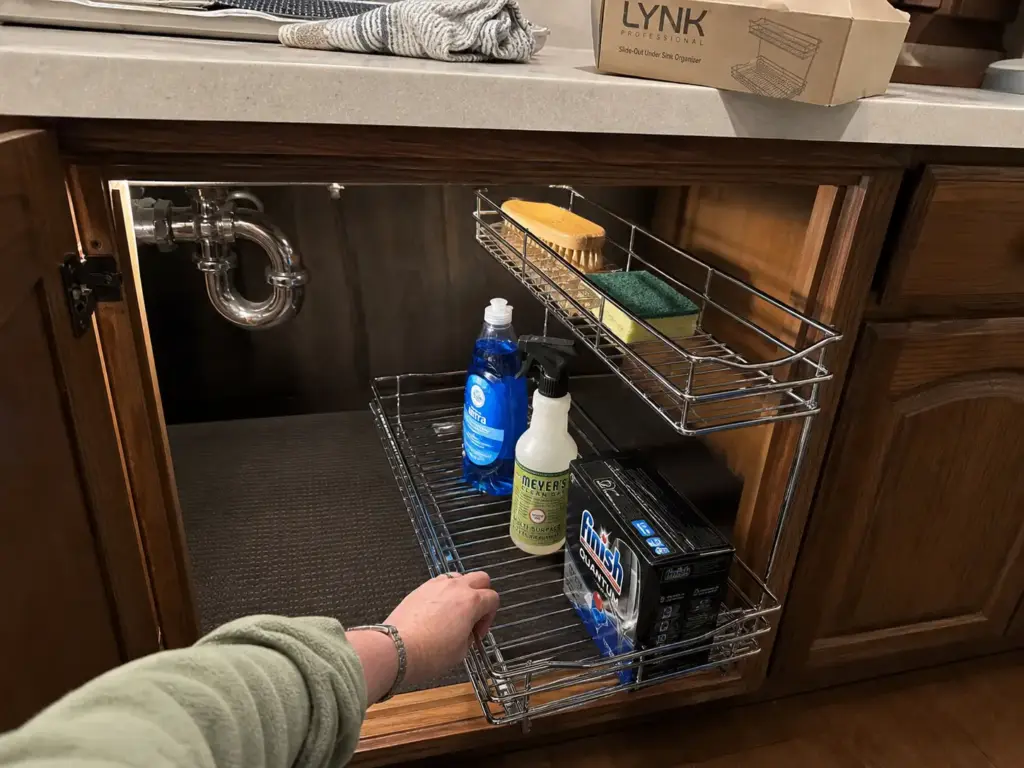Hand sliding out a LYNK pull-out under sink organizer mounted beside an off-center kitchen drain