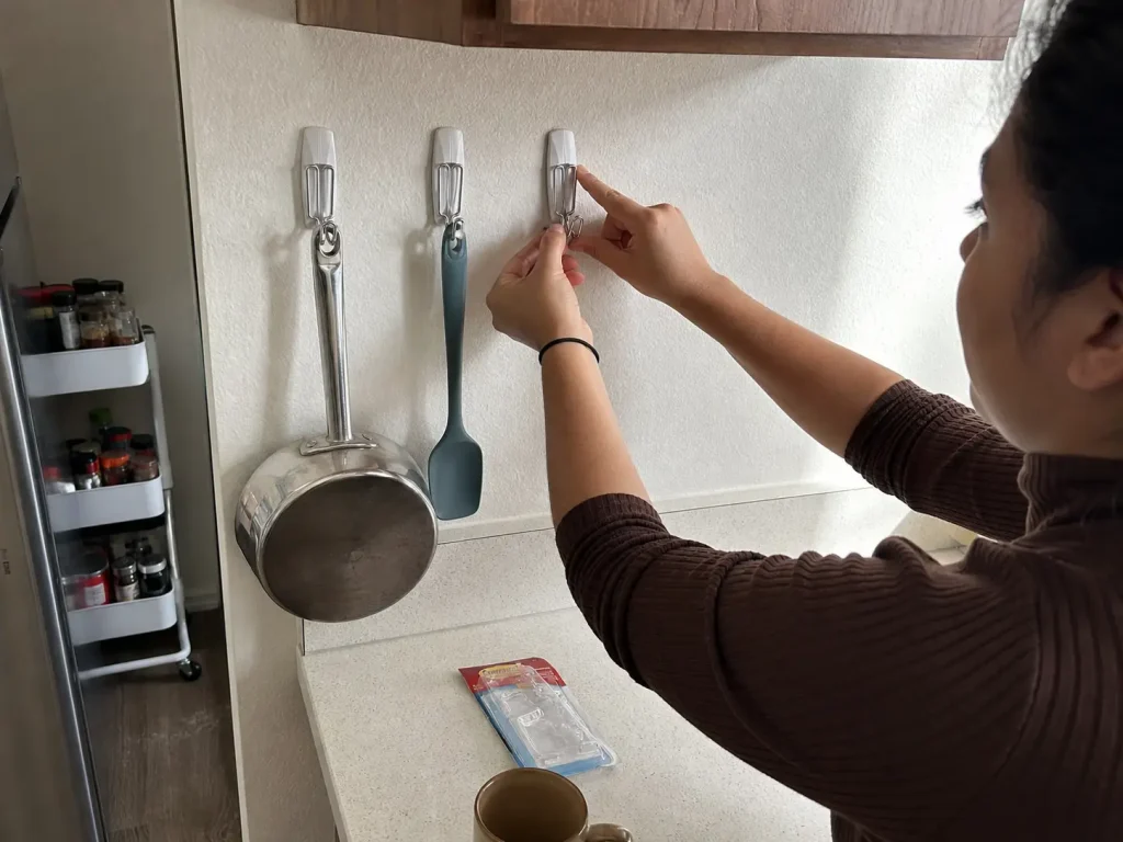 Why Your Small Kitchen With Few Drawers Stays Cluttered 6 Hands pressing an adhesive wire hook onto a rental kitchen wall to hang light utensils without drilling