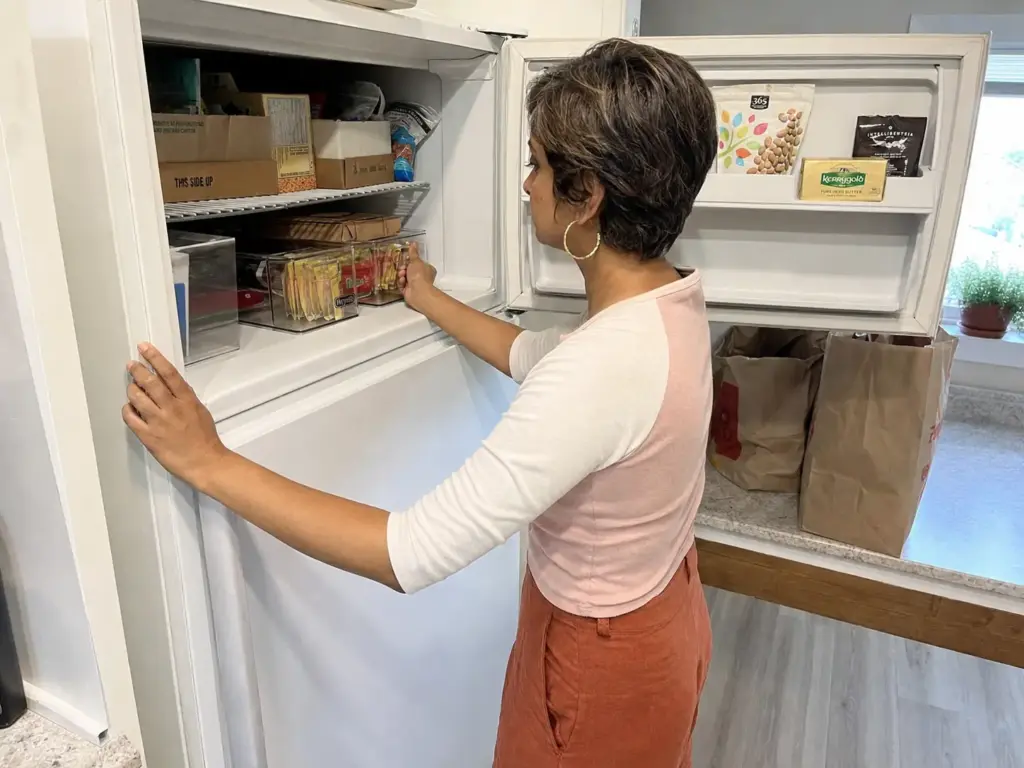 Renter sliding a low clear bin under the single shelf of a small over-fridge freezer to add a tier