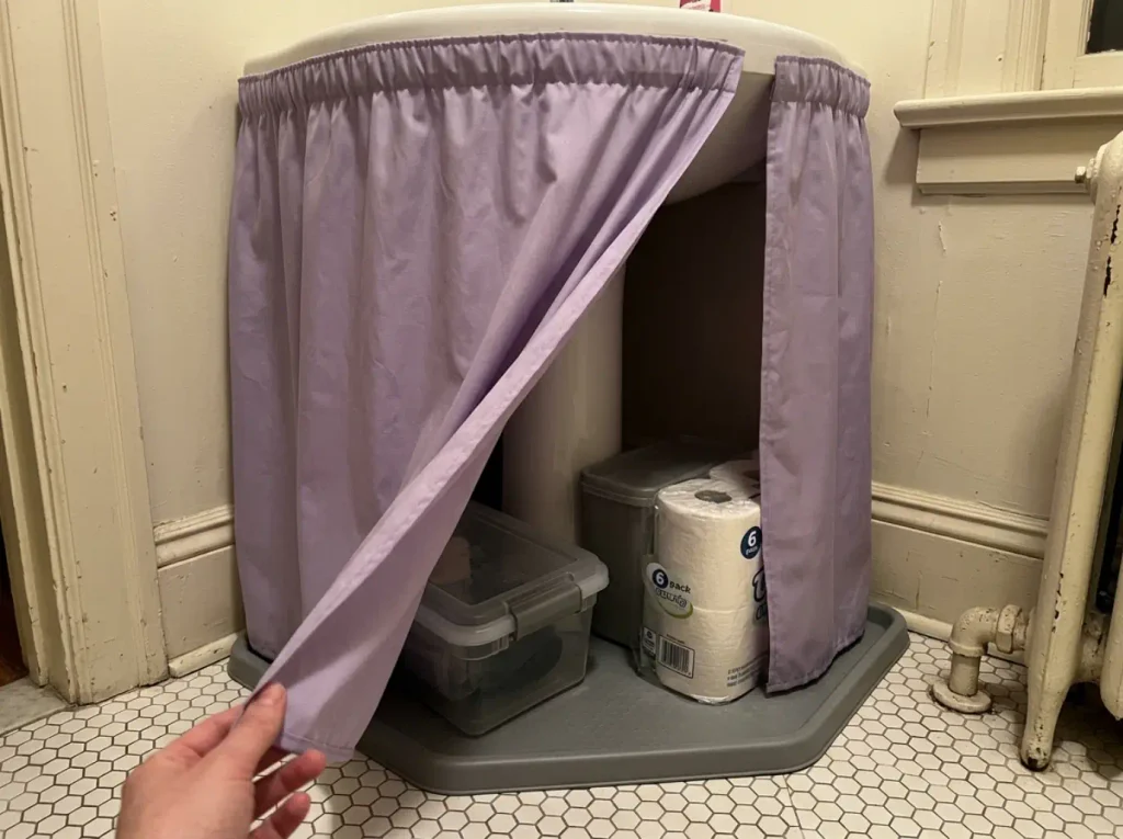 Fabric sink skirt hanging around a pedestal sink with one edge lifted showing bins stored on the floor