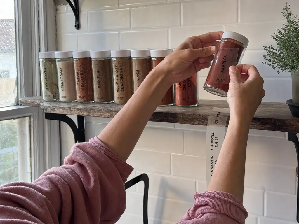 Hands placing a clear spice jar label on a glass jar with matching jars on an open shelf