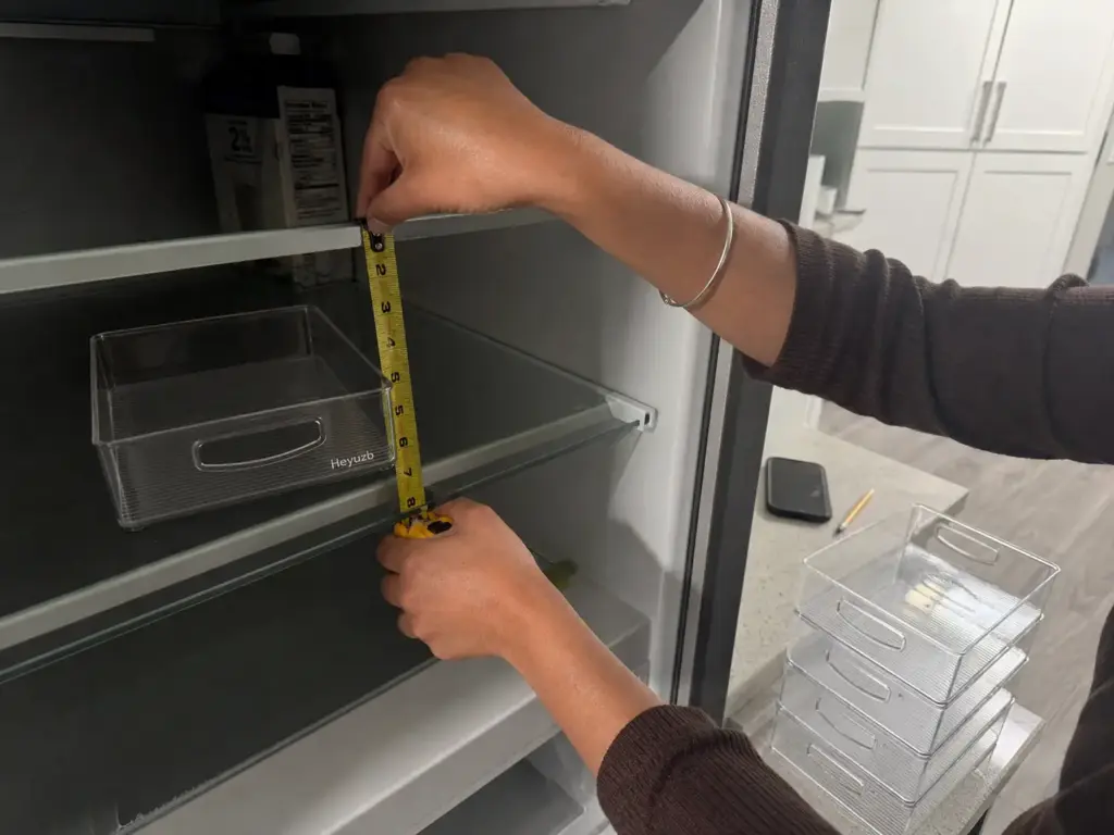 Hands measuring fridge shelf clearance with a tape measure next to a clear stackable organizer bin