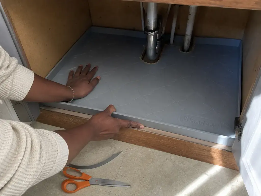 Hands laying a trimmed waterproof under sink mat with a raised lip around the kitchen drain pipes