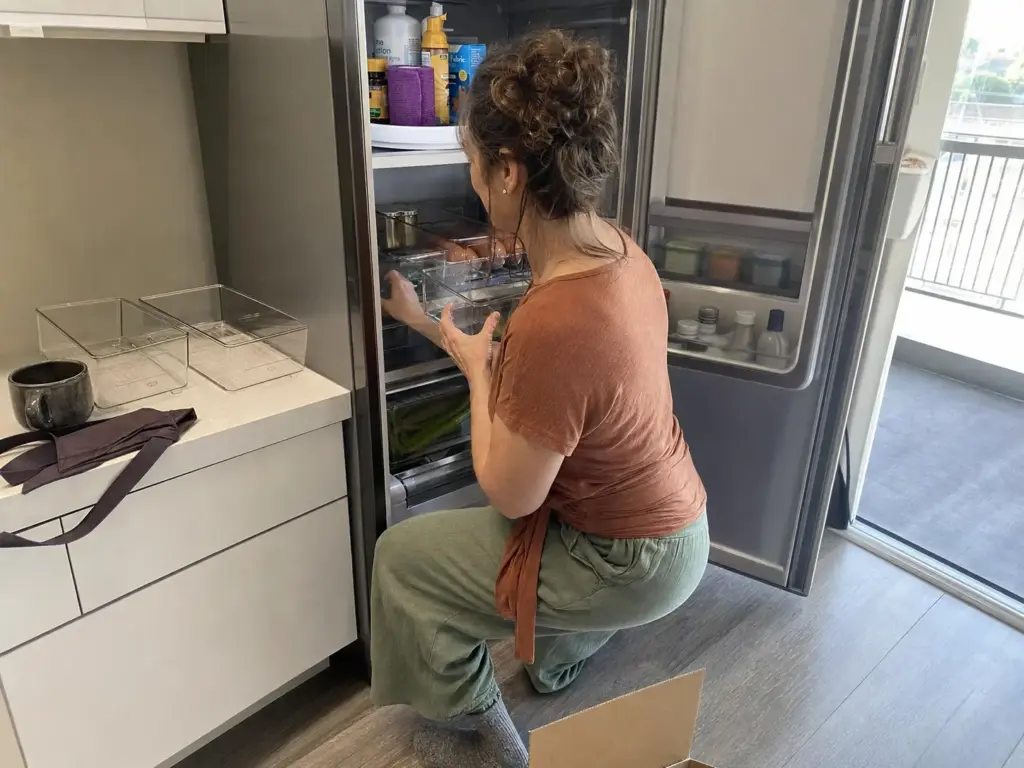 Woman placing clear fridge bins and a small turntable of condiments onto a refrigerator shelf