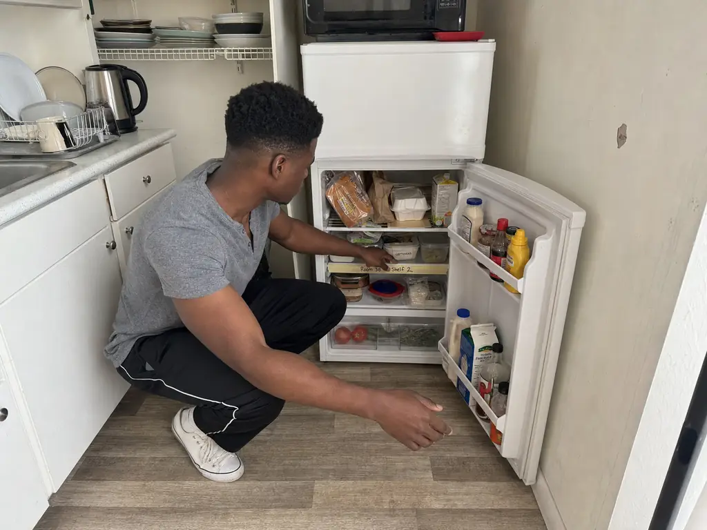 Person crouching in front of a compact apartment refrigerator with one shelf organized and the rest shared