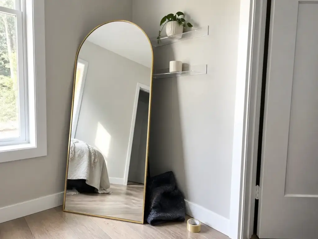 Full length arched mirror leaned against a small bedroom wall bouncing window light