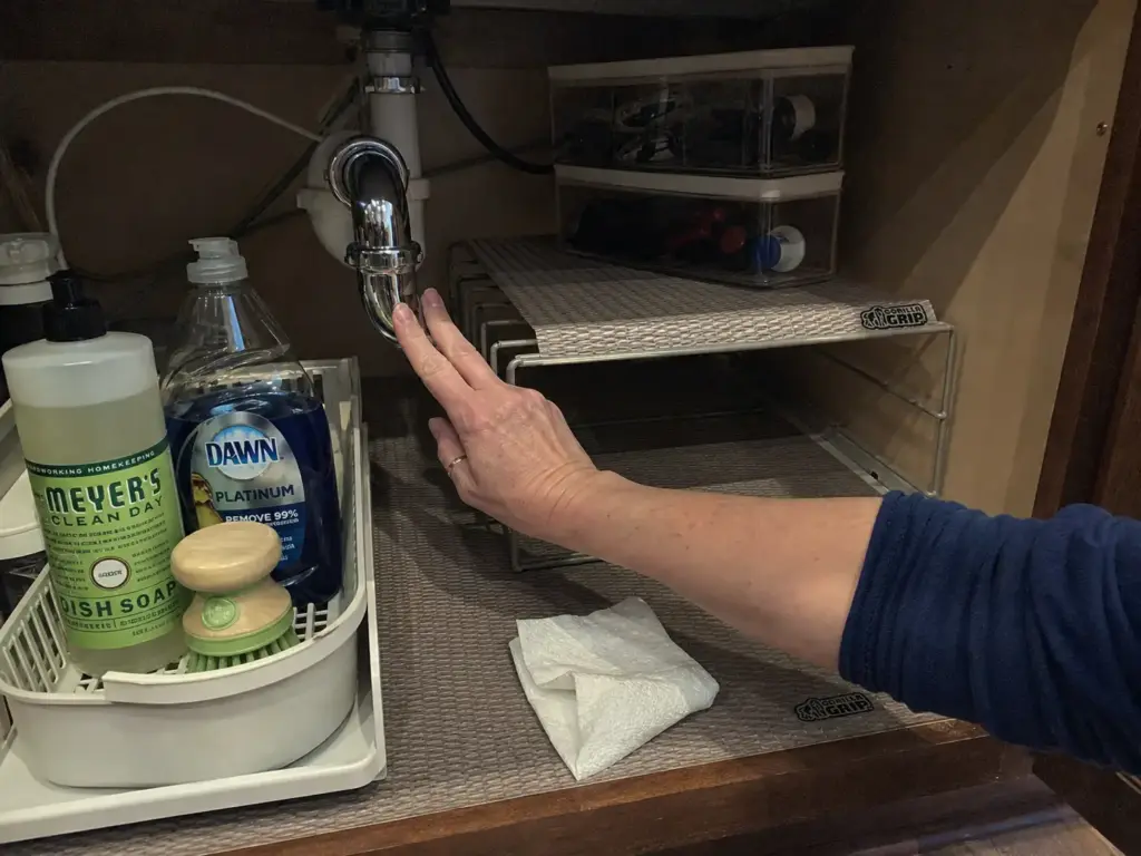 Hand checking under the kitchen sink P-trap for dampness during a monthly under sink reset
