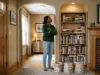 7 Bookshelf Styling Ideas That Survive Real Life Woman standing back from a half-styled bookcase in an arched alcove, books stacked on the rug beside her