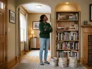 7 Bookshelf Styling Ideas That Survive Real Life Woman standing back from a half-styled bookcase in an arched alcove, books stacked on the rug beside her