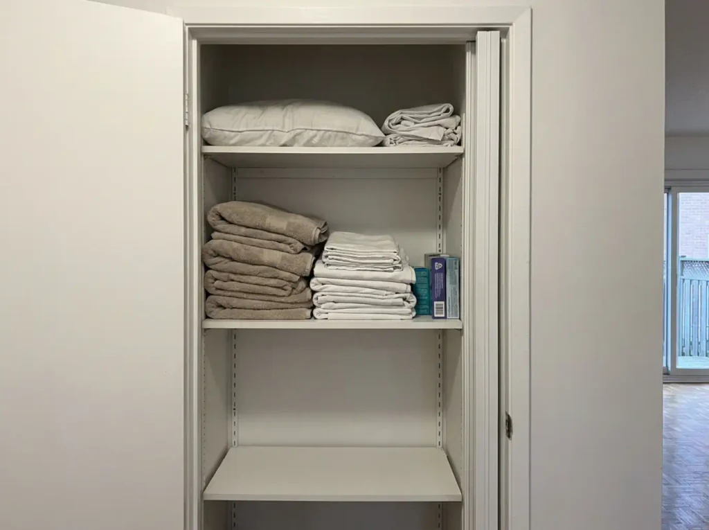 Cluttered hallway linen closet with mixed towels, sheets and toiletry backstock on one shelf