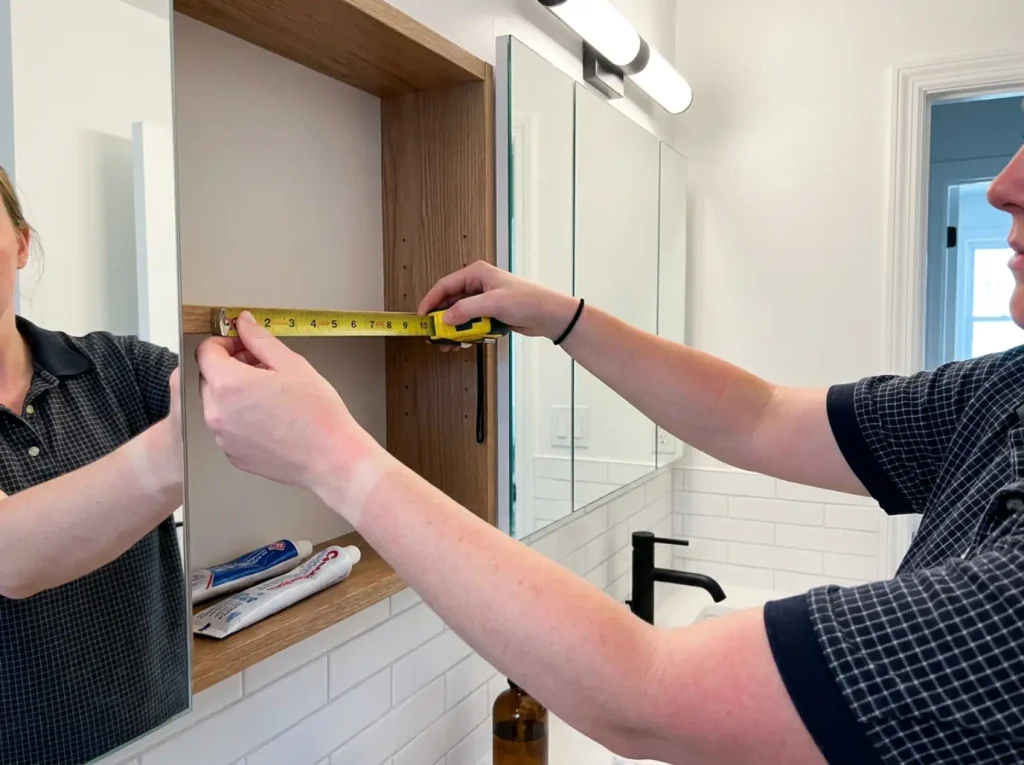 Hands measuring the shallow shelf depth of an open recessed medicine cabinet with a tape measure